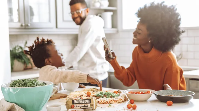 A family laughing together as they make pizza at the kitchen counter, one of them capturing the moment on a phone