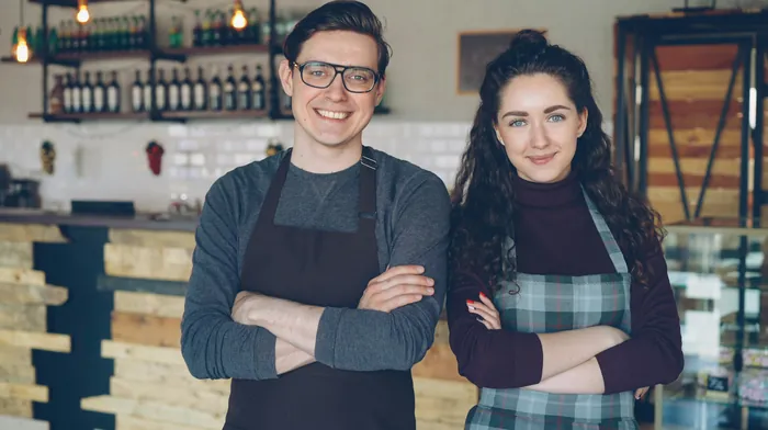 Two café owners in aprons standing proudly in front of their counter