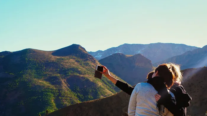 Two friends hugging on a mountainside while one photographs the view on her phone
