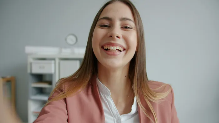 A smiling young woman in a pink blazer speaking straight to the camera